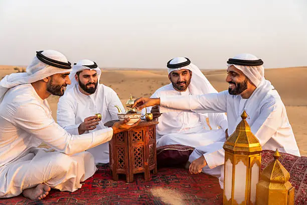 Four Emirati men in traditional white kandura sitting on carpet in desert sharing Arabic coffee and dates during authentic Dubai desert safari cultural experience