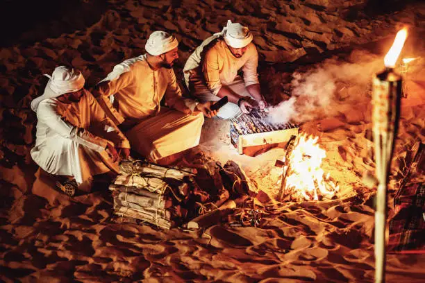 Three Arab men in traditional clothing grilling kebabs over open campfire on desert sand dunes at night during authentic Dubai evening desert safari BBQ experience