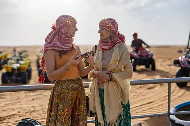 Two happy female tourists wearing traditional Arabic headscarves and colorful outfits chatting near quad bikes on desert sand dunes during Dubai desert safari adventure