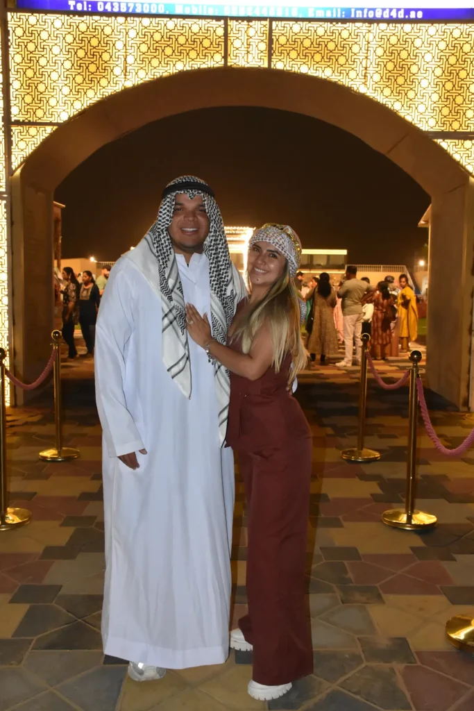 Tourist couple dressed in traditional Emirati kandura and headscarf at a Dubai desert safari camp entrance at night